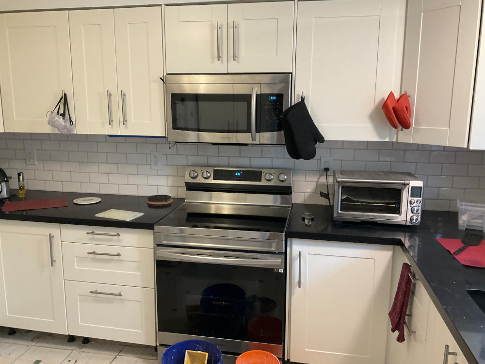 photo of my almost-finished kitchen with the white cabinets, brushed nickel door handles, an over-the-range microwave, black quartz countertops, range, and smaller countertop appliances with the newly installed white subway tile with warm grey grouting